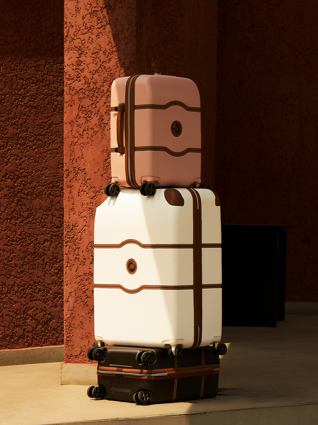Delsey koffers uit de Iconische serie Chatelet Air 2.0, een roze koffer, een witte koffer en een bruine koffer voor een terracotta kleurige muur.